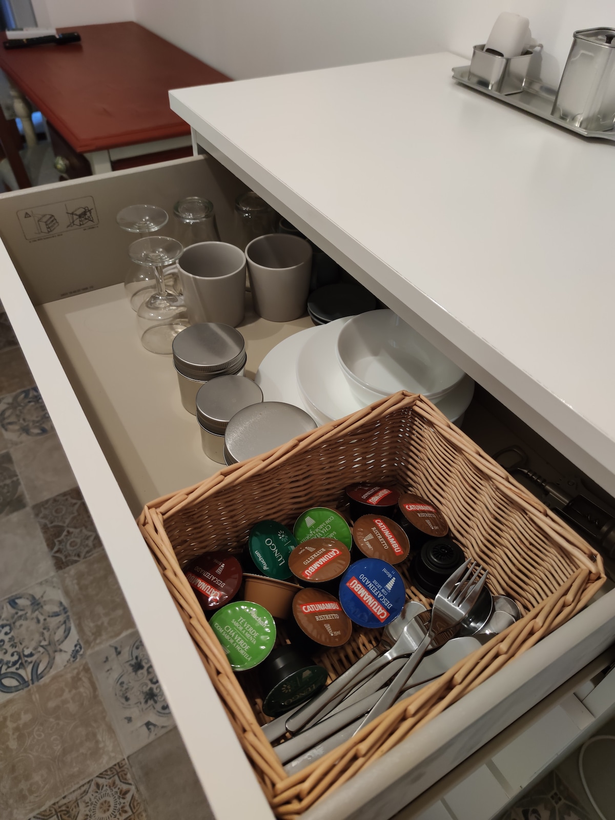 A kitchen drawer displays glassware, cups, and plates organized neatly. A woven basket contains various coffee capsules in assorted colors, along with utensils. The drawer incorporates a clean and modern design, emphasizing practicality for guest use.