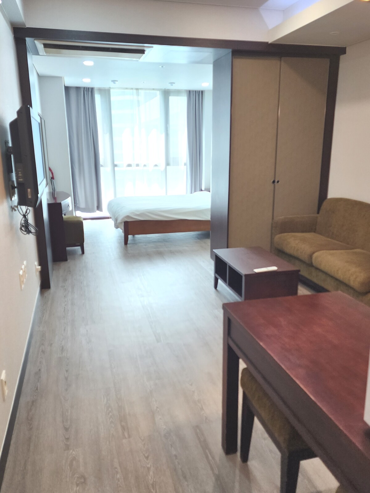 A bright and airy guest room is featured, with a spacious bed positioned against the wall. A small table and two chairs are visible, along with a cozy seating area. Large windows allow natural light to fill the space, complemented by neutral-colored furnishings.