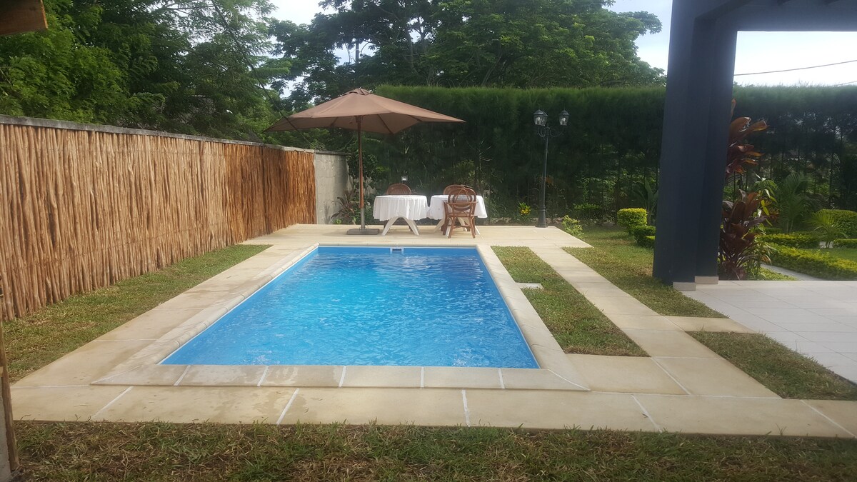A private pool is surrounded by well-maintained grass and a stone patio. An umbrella shades a small table and two wooden chairs nearby, creating a relaxing spot. Lush greenery is seen in the background, adding to the serene ambiance of the outdoor space.