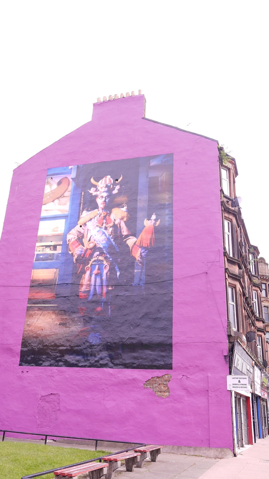 A large mural on a purple wall displays an artistic figure, contributing to the vibrant street scene. The mural's background contains hints of a bustling exterior, while nearby benches provide a place for visitors to relax. The top of the building features a chimney.