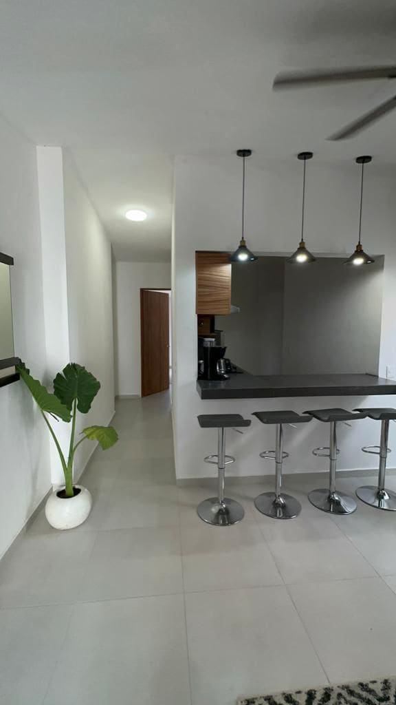 A modern kitchen area features a sleek countertop with four metal bar stools positioned underneath. Pendant lights hang above, illuminating the space. A tall indoor plant adds a touch of greenery, while a mirrored wall reflects the minimalistic design.