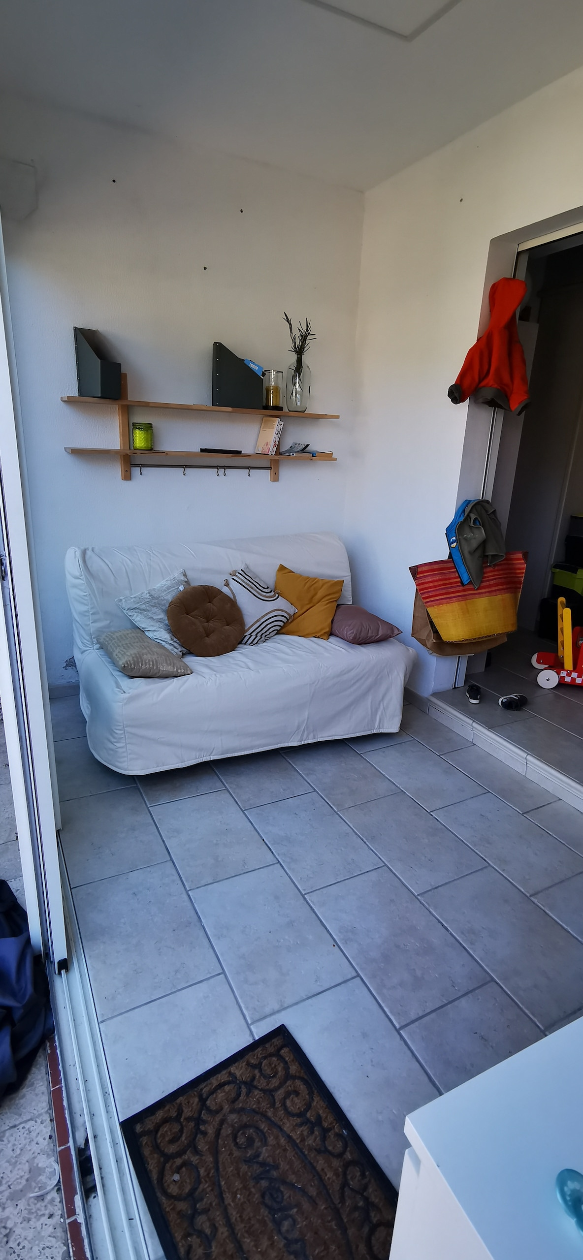 A small sitting area features a white couch adorned with cushions in various colors. A shelf above the couch displays decorative items, while a corridor leads to another room. A door opens to an outdoor space, enhancing a bright and airy feel.