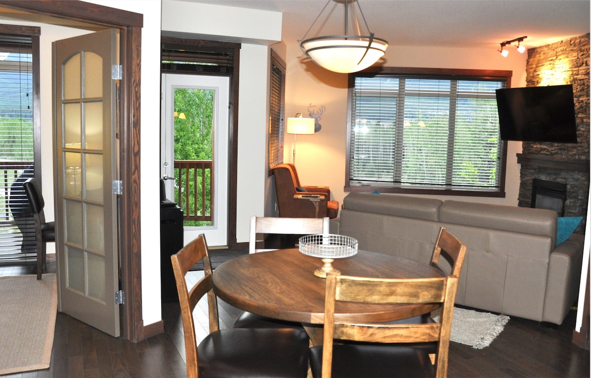 An open layout is displayed, featuring a dining table with four wooden chairs, leading to a comfortable living area with a large beige sofa. Natural light enters through multiple windows, creating a welcoming atmosphere. A stone fireplace highlights one corner, enhancing the cozy design.