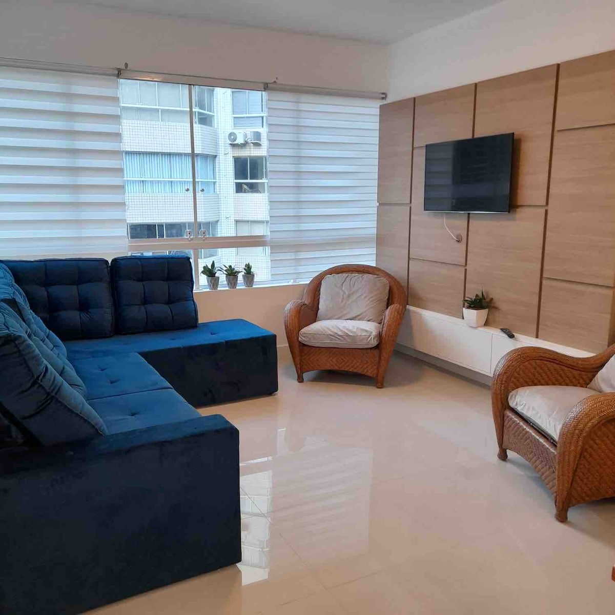 A modern living area features a blue sectional sofa paired with two wicker chairs. A wall-mounted television is visible, along with large windows dressed in sheer shades, allowing natural light to fill the room. Decorative plants add a touch of greenery.