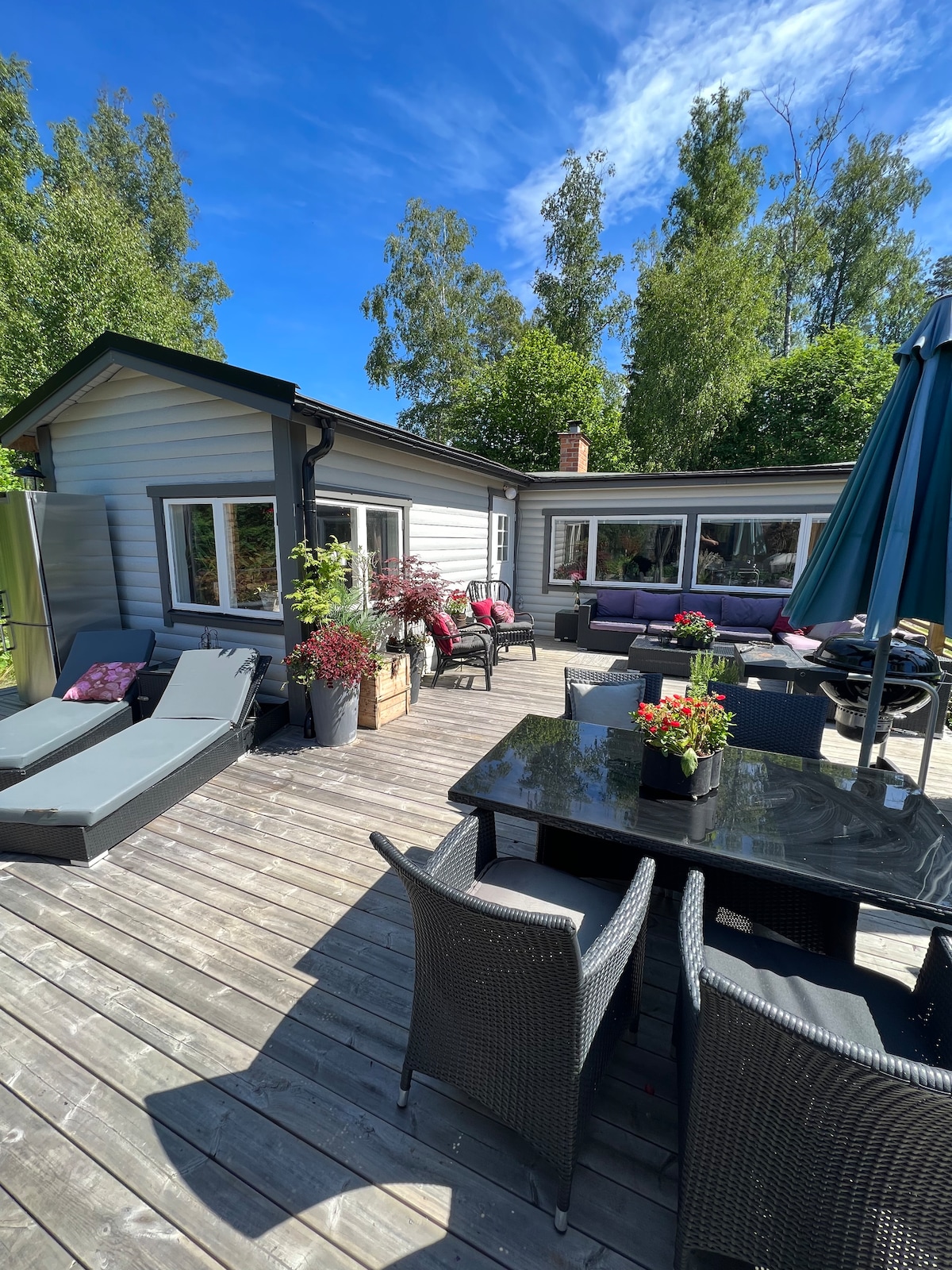 A wooden deck is showcased, featuring a dining table surrounded by several black wicker chairs. Sun loungers in muted tones sit adjacent, complemented by potted plants and vibrant flowers. A shaded area with an umbrella is visible, providing comfort in sunny weather.