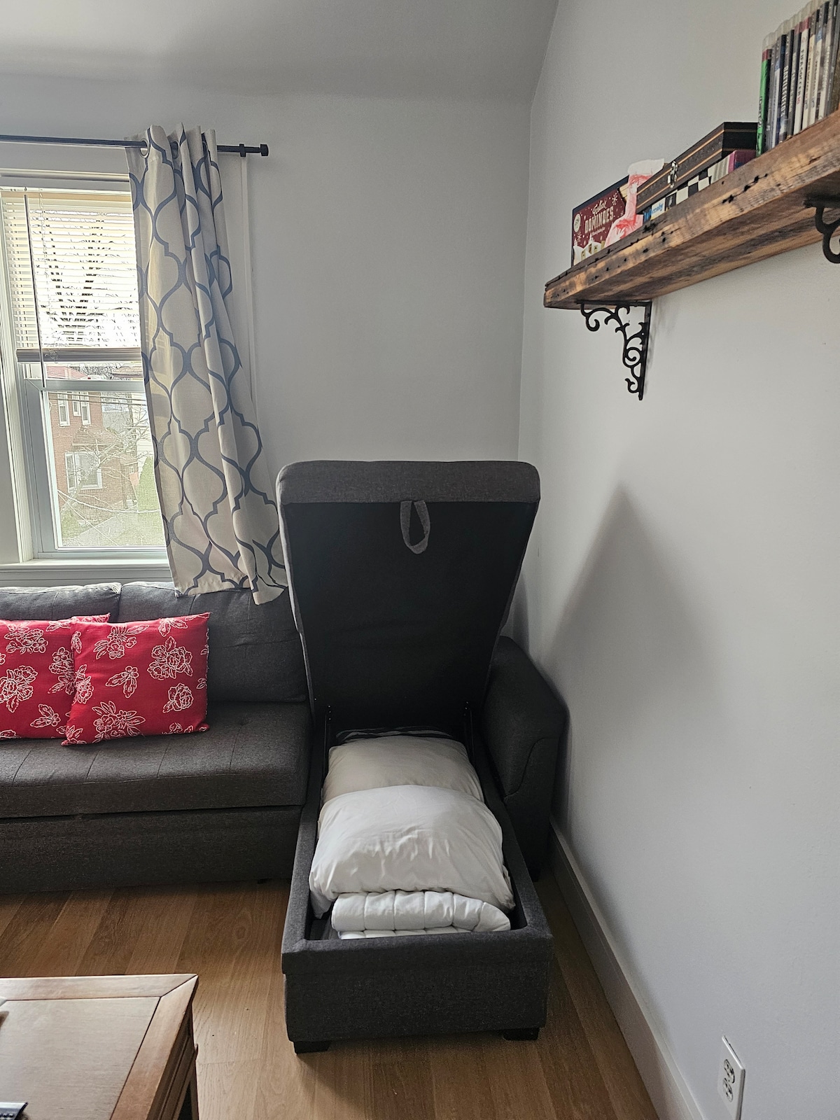 A cozy living room corner displays a gray sofa with decorative red and white cushions. A nearby window allows natural light to enter, illuminating a storage ottoman that is partially open, revealing soft bedding inside. A wooden shelf holds various items above.