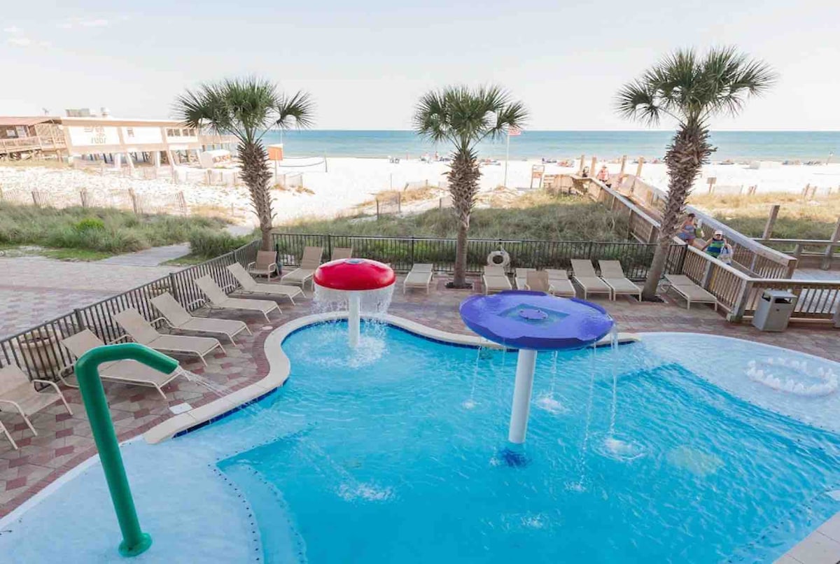 The pool area features a colorful design with a blue swimming pool accentuated by two vibrant water features in red and purple. Sun loungers are arranged around the pool, with palm trees providing a tropical ambiance. The beach is visible in the background.