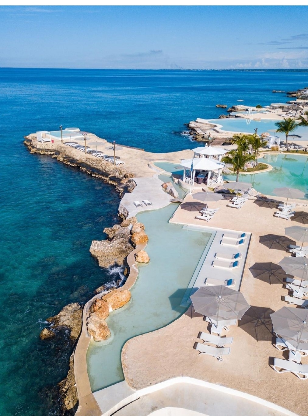 An aerial view captures a serene coastal scene, featuring clear turquoise waters meeting a rocky shoreline. Lounge chairs and umbrellas line a spacious patio area, providing ample seating. The smooth curves of the pool extend towards the ocean, creating a tranquil environment perfect for relaxation.