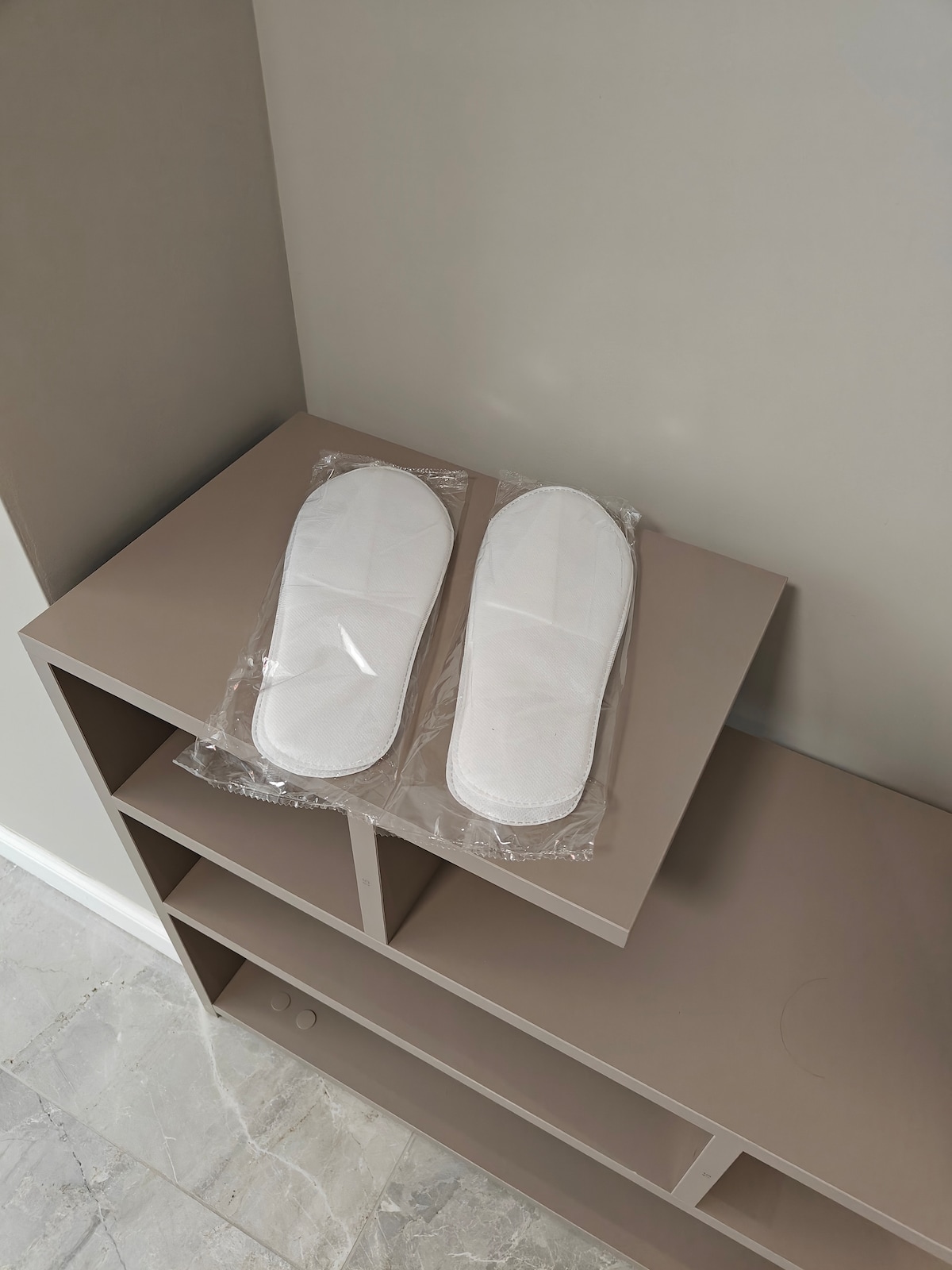 Two pairs of wrapped white slippers are displayed on a beige shelf. The shelf features a minimalist design with open compartments beneath. The light-colored flooring enhances the neutral tones of the decor.