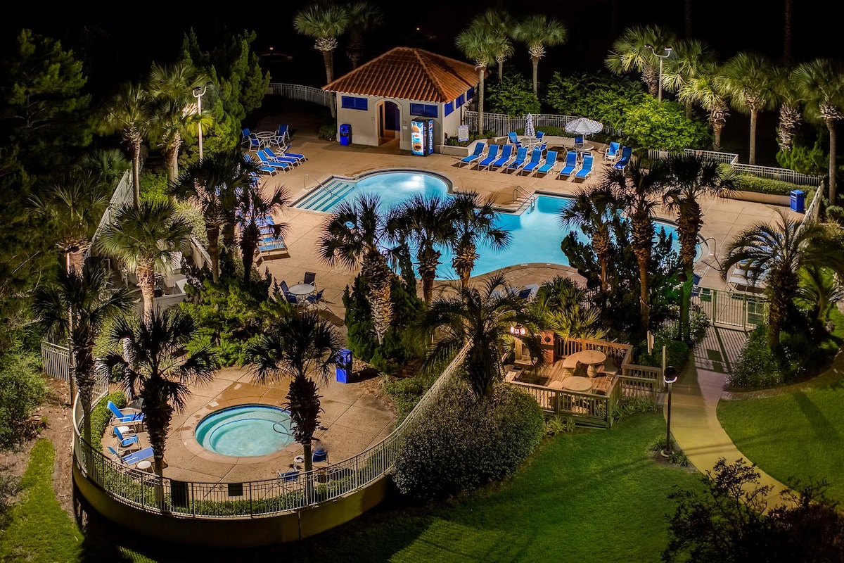 An aerial view reveals a well-kept pool area surrounded by lush palm trees. A hot tub is situated near the edge, with loungers evenly spaced around the larger pool. A shaded building is visible, providing restroom facilities and refreshment options. The scene is illuminated, enhancing its inviting atmosphere.