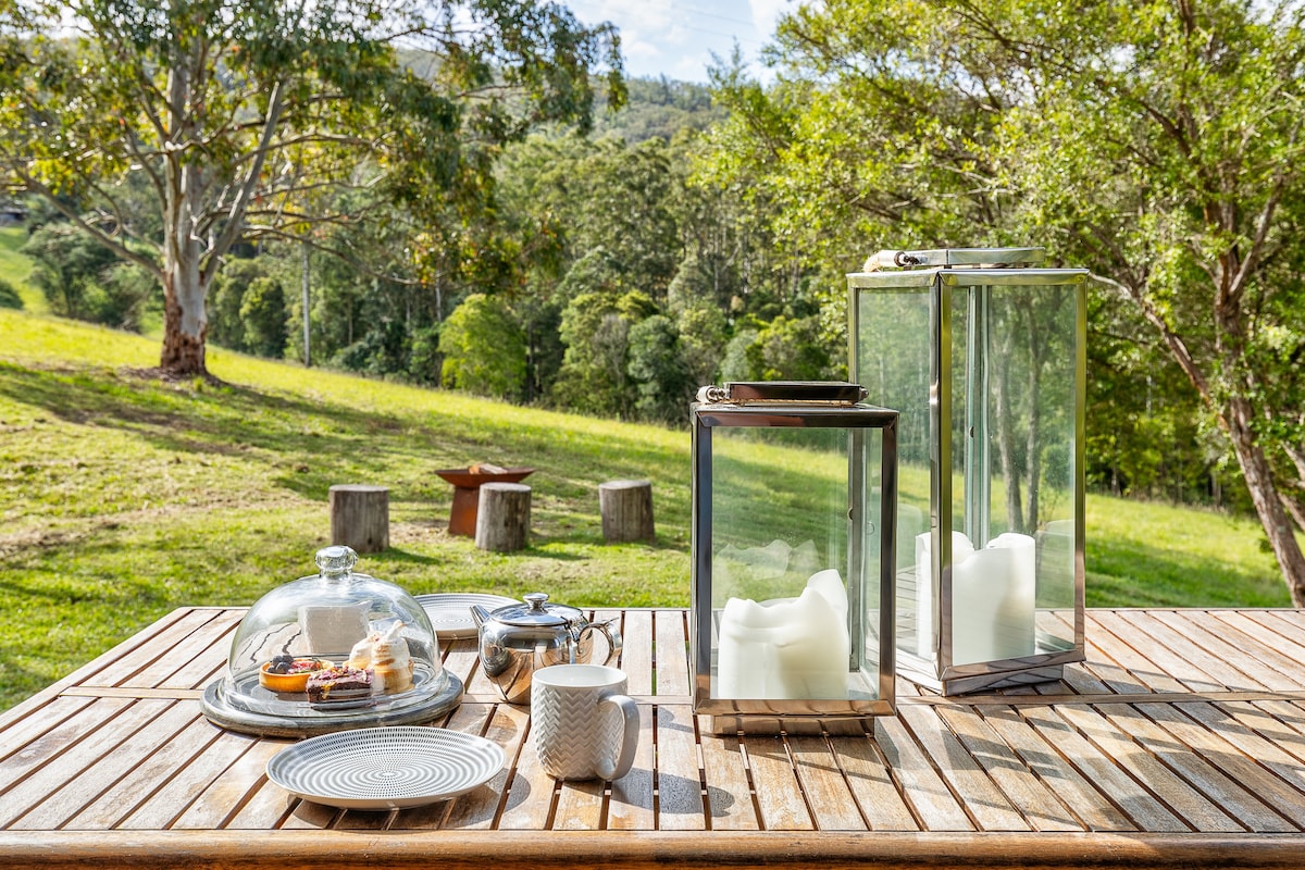A wooden outdoor table is set with a glass cake dome containing baked goods, accompanied by a teapot and cups. Two lanterns hold glowing candles, and lush greenery is visible in the background, showcasing a scenic view of the surrounding landscape.