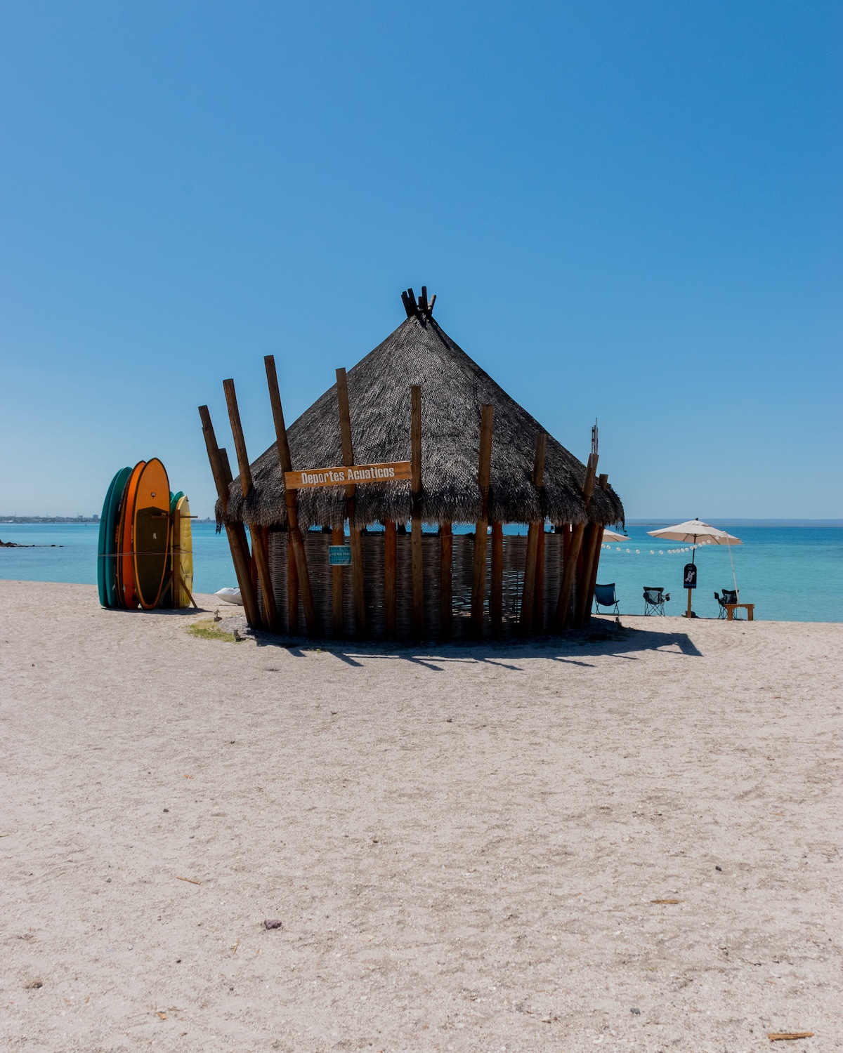 A beach hut with a thatched roof stands on a sandy shoreline, surrounded by clear blue waters. Nearby, colorful surfboards lean against a post. A shaded seating area with an umbrella offers a place to relax and enjoy the coastal view.