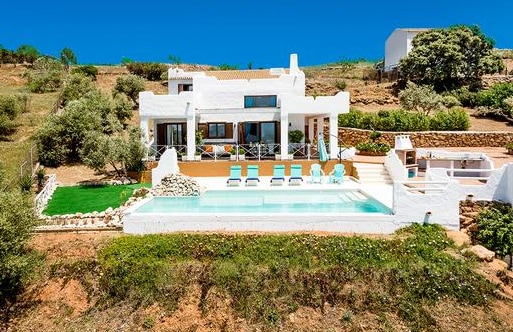 A modern white villa is set against a hillside, featuring a spacious terrace with a swimming pool. Lounge chairs are arranged alongside the pool, and well-maintained greenery surrounds the property. The design incorporates natural elements and large windows that invite outdoor light.