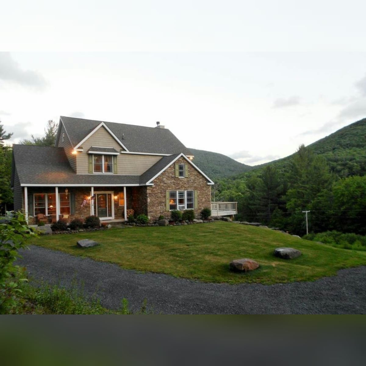 This spacious home is set against a backdrop of rolling mountains, featuring a combination of stone and wood siding. Outdoor landscaping includes a manicured lawn and natural stone accents, with a gravel driveway providing access. The sky dims gently, suggesting the approach of evening.