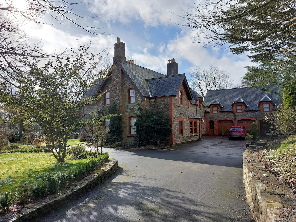 The exterior of a spacious house is showcased, featuring traditional architecture with a combination of stone and brick elements. A large driveway leads up to the entrance, surrounded by neatly trimmed gardens and a variety of trees. Parking spaces are visible, with a red vehicle parked nearby.
