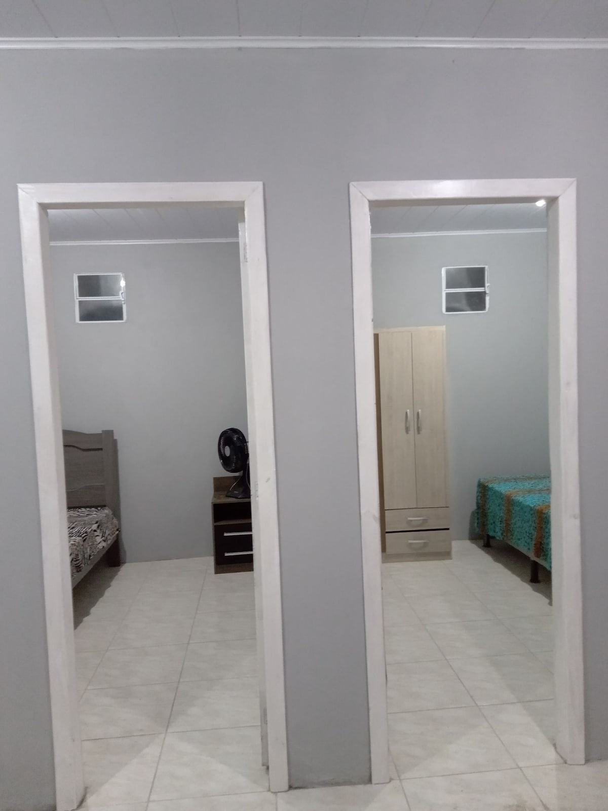 Two separate rooms are visible, each featuring distinct beds and storage options. One room contains a fan, while the other showcases a chest of drawers. Neutral grey walls and tile flooring create a calm environment, enhanced by natural light entering through small windows.