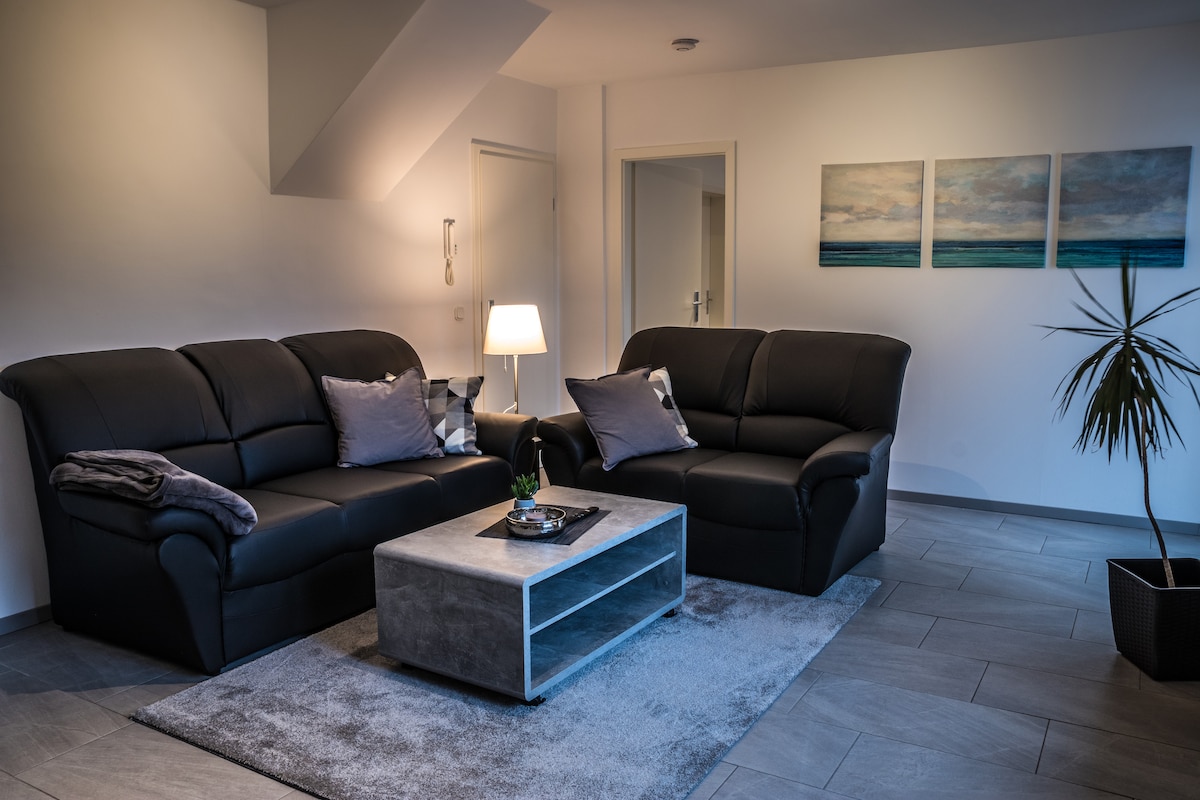 A living area features two black leather sofas, adorned with cushions, positioned around a modern coffee table. A soft carpet lies beneath, and a lamp casts a warm glow, enhancing the space. Three abstract artworks hang on the wall, complementing the neutral tones.
