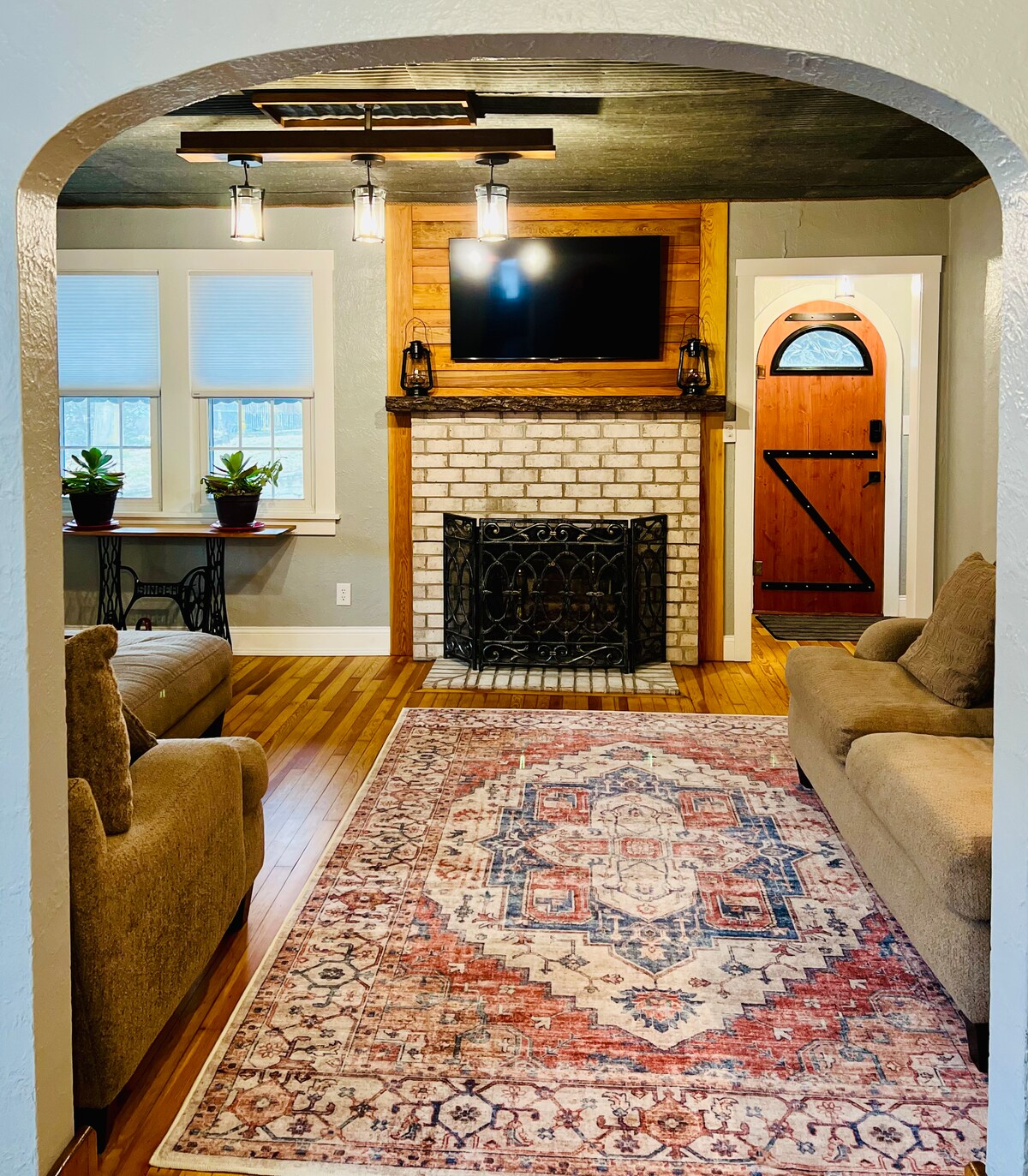 A cozy living room is displayed, featuring a decorative fireplace with a dark metal screen. Two comfortable sofas are positioned on either side of a colorful area rug. A flat-screen TV is mounted above the fireplace, and plants are placed on the window sill.