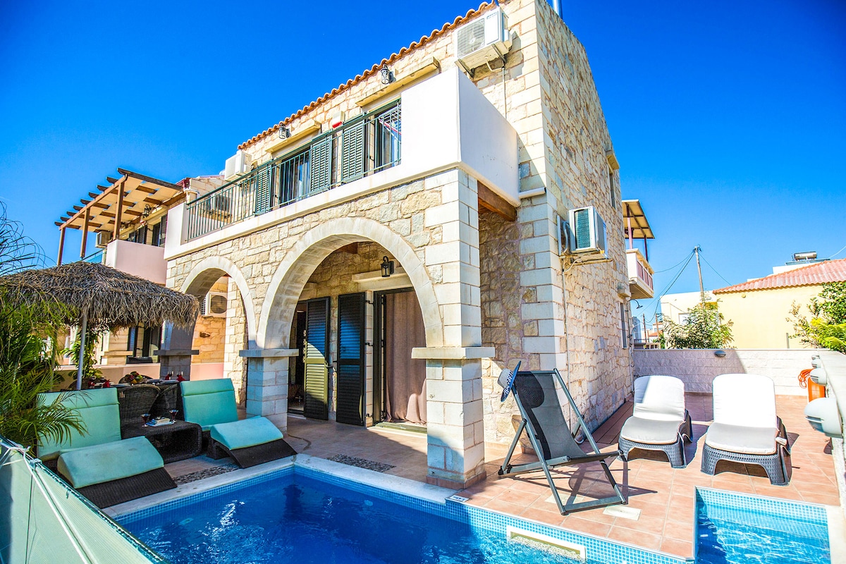 A two-story villa features a combination of stone and modern architecture, creating a charming exterior. A private swimming pool is visible in the front, surrounded by sun loungers and shaded sitting areas. Balconies offer views of the clear blue sky and surrounding greenery.