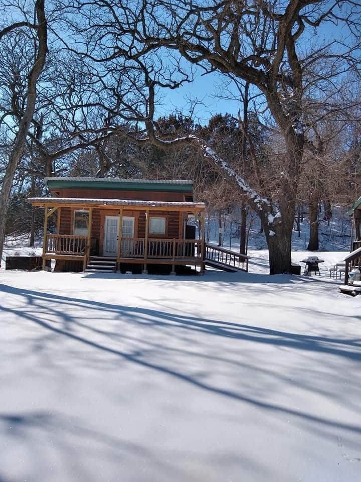 A quaint cottage is nestled among snow-covered trees, surrounded by a blanket of fresh snow. The porch, with wooden railing, provides access to the entrance. A grill is situated nearby, ready for outdoor cooking, under a clear blue sky.