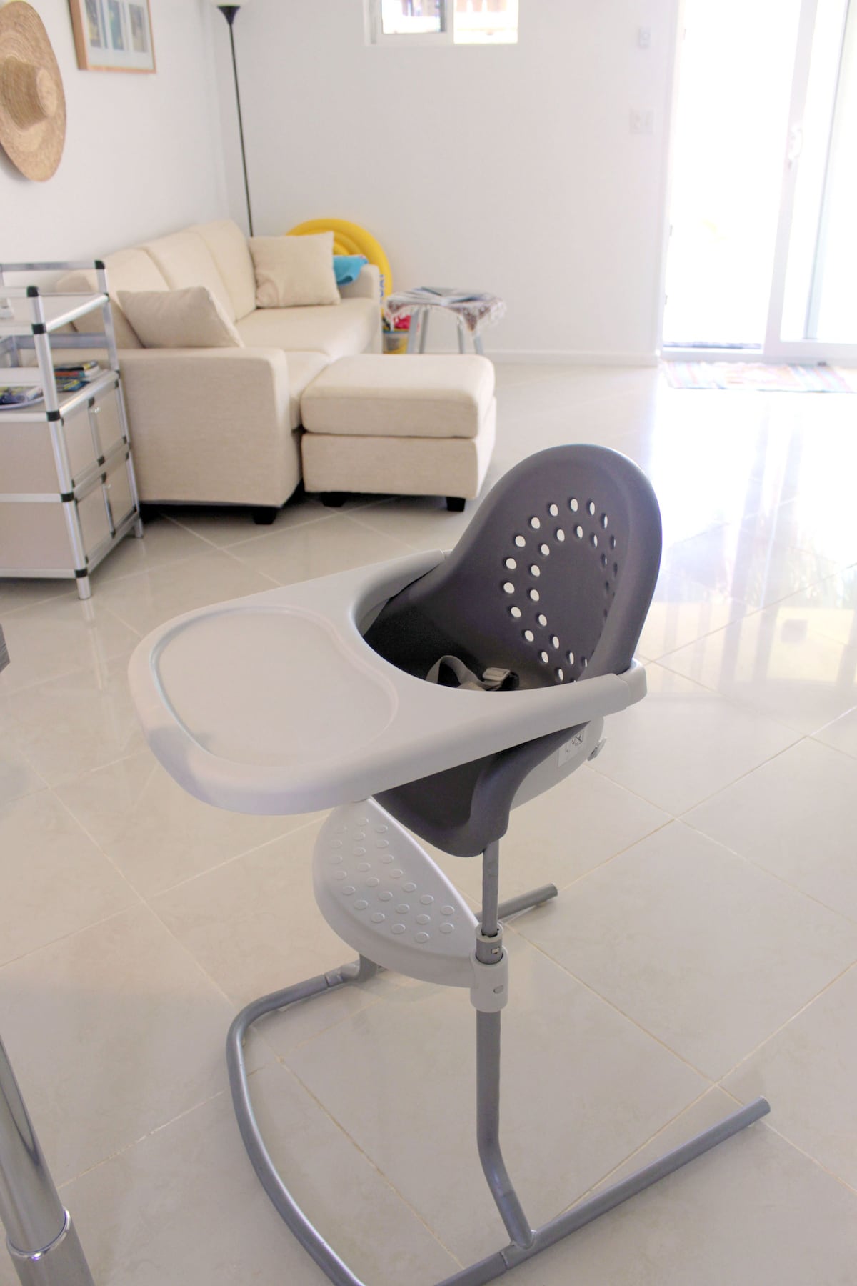 A modern high chair is positioned in a bright living area, featuring a sleek design with a safety tray and ventilation holes. Light enters through large glass doors, illuminating the nearby neutral-colored sofa and inviting atmosphere of the space.