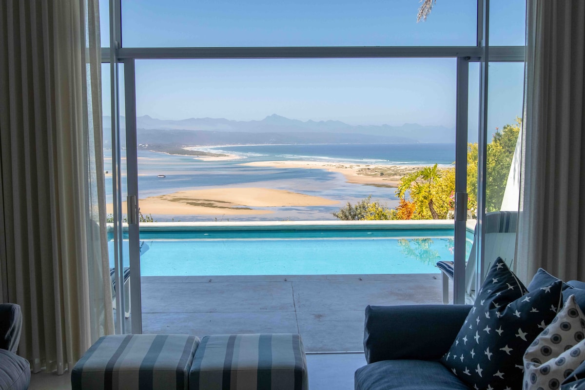 A spacious seating area features comfortable armchairs and a footrest, positioned to overlook an expansive view of the ocean, lagoon, and distant mountains. Large glass panels allow abundant natural light, emphasizing the seamless connection to the outdoor pool and scenic landscape.