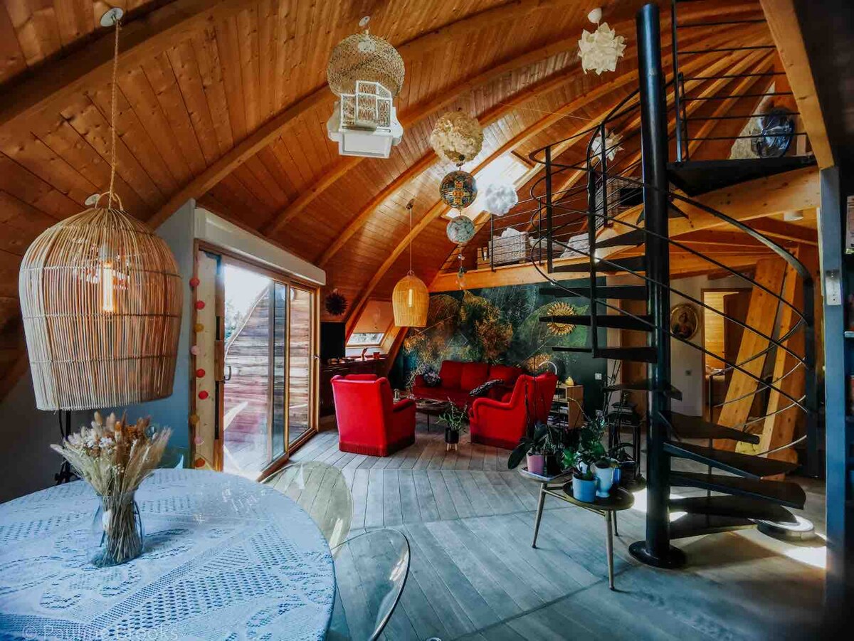 The interior features a spiral staircase leading to an upper level, surrounded by wooden beams and natural light. A dining table sits nearby, adorned with a lace tablecloth, while red seating areas create a cozy space, enhanced by decorative elements and greenery.