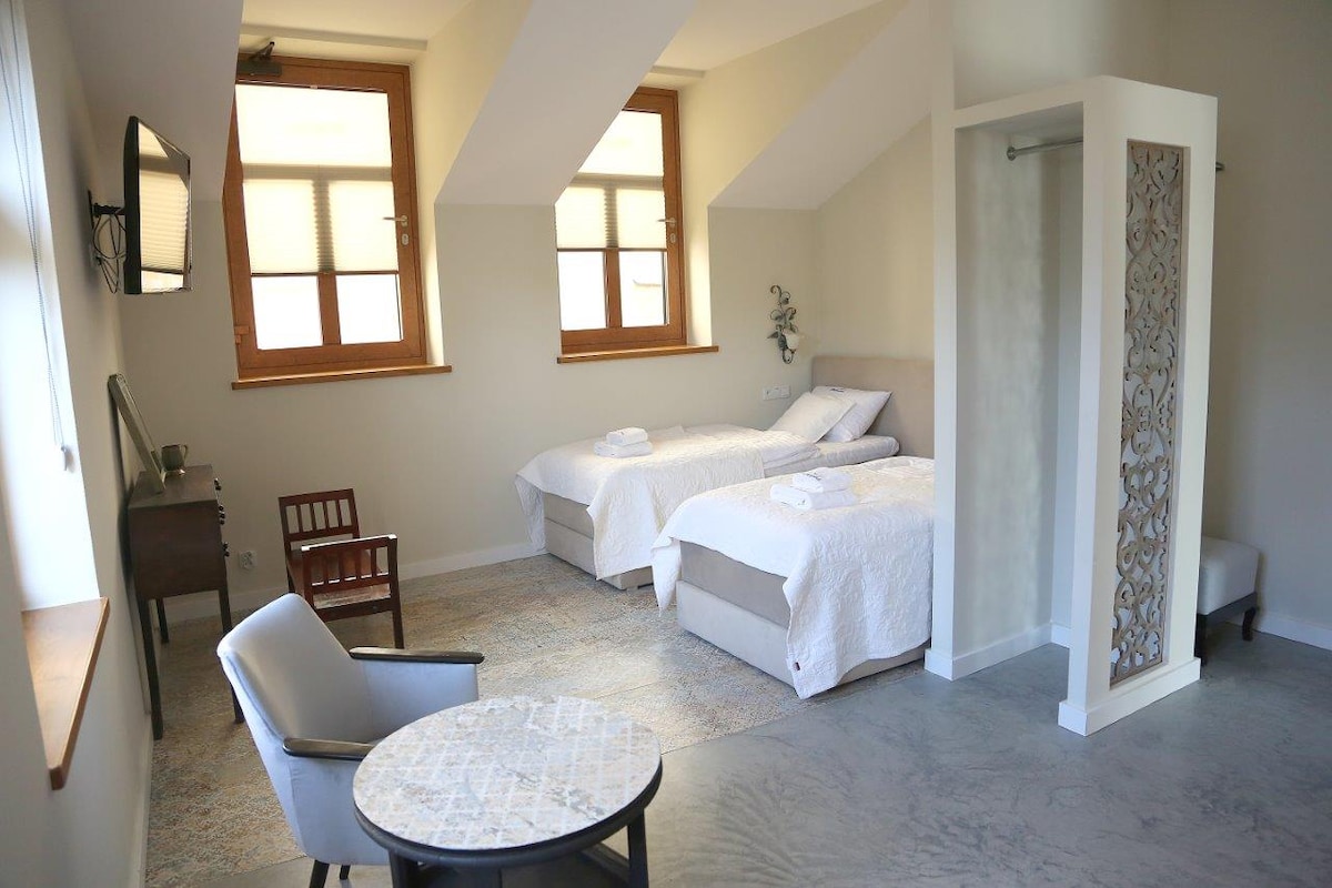 A light-filled room features two spacious beds with fresh linens arranged side by side. A small table and chair provide a cozy seating area, while a glass-enclosed shower is present in the corner. Large windows allow natural light to brighten the space.