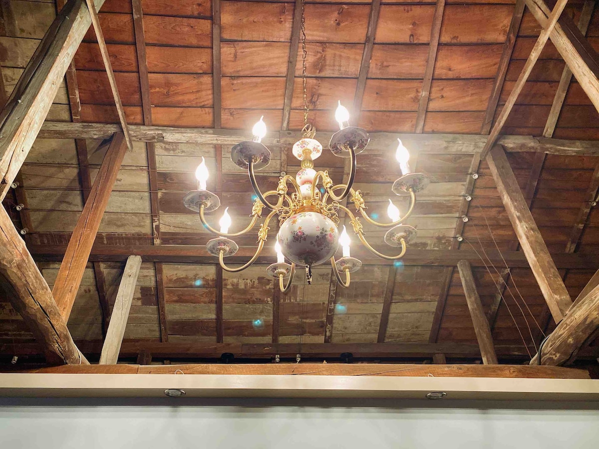 An ornate chandelier hangs from a wooden ceiling, showcasing intricate detailing and multiple light bulbs illuminating the space. Exposed wooden beams add a rustic charm, contrasting with the elegant design of the fixture above.