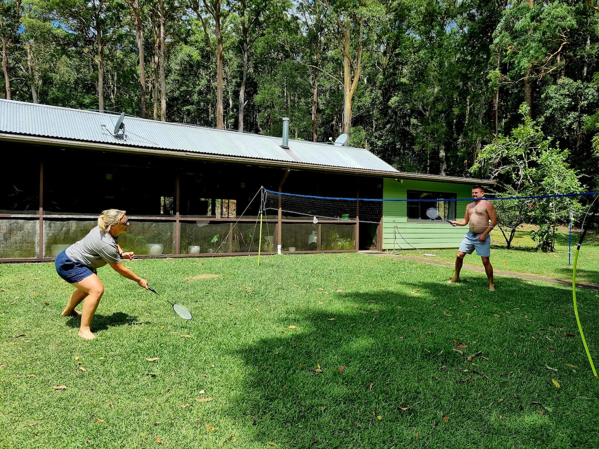 A grassy backyard is depicted, where two individuals engage in a casual game of badminton. The house is visible in the background, featuring a slanted roof and large windows. Surrounding trees provide a natural backdrop, contributing to the private and serene atmosphere.