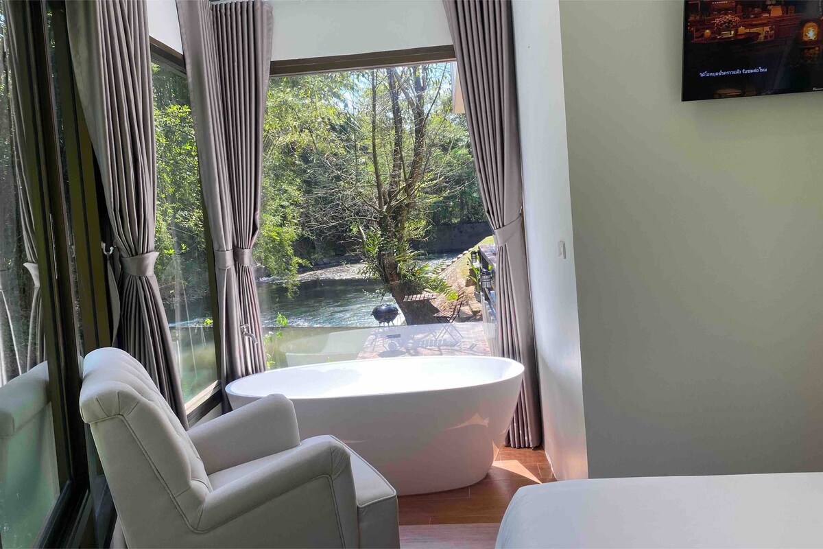A well-lit room features a freestanding bathtub positioned near large windows, offering views of a tranquil stream and greenery outside. Gray drapes frame the windows, complementing the simple chair and minimalist decor in the space.