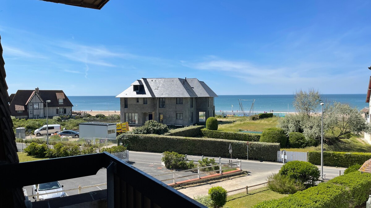 A view from the balcony captures the serene coastline and beach, with a sandy shore stretching into the distance. The image showcases a charming building across the street surrounded by greenery, providing a tranquil atmosphere.