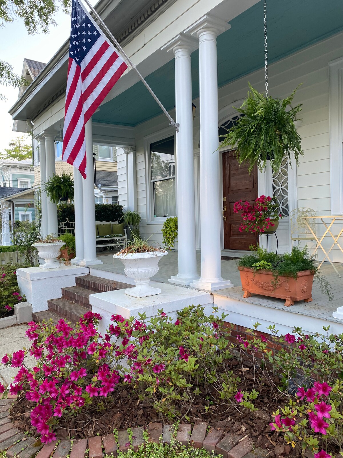 A charming porch is highlighted by white columns and an American flag, creating an inviting entrance. Lush hanging ferns and colorful planters with blooming flowers add to the warm ambiance, while the laid-brick pathway leads visitors towards the entrance, enhancing a sense of comfort and welcome.