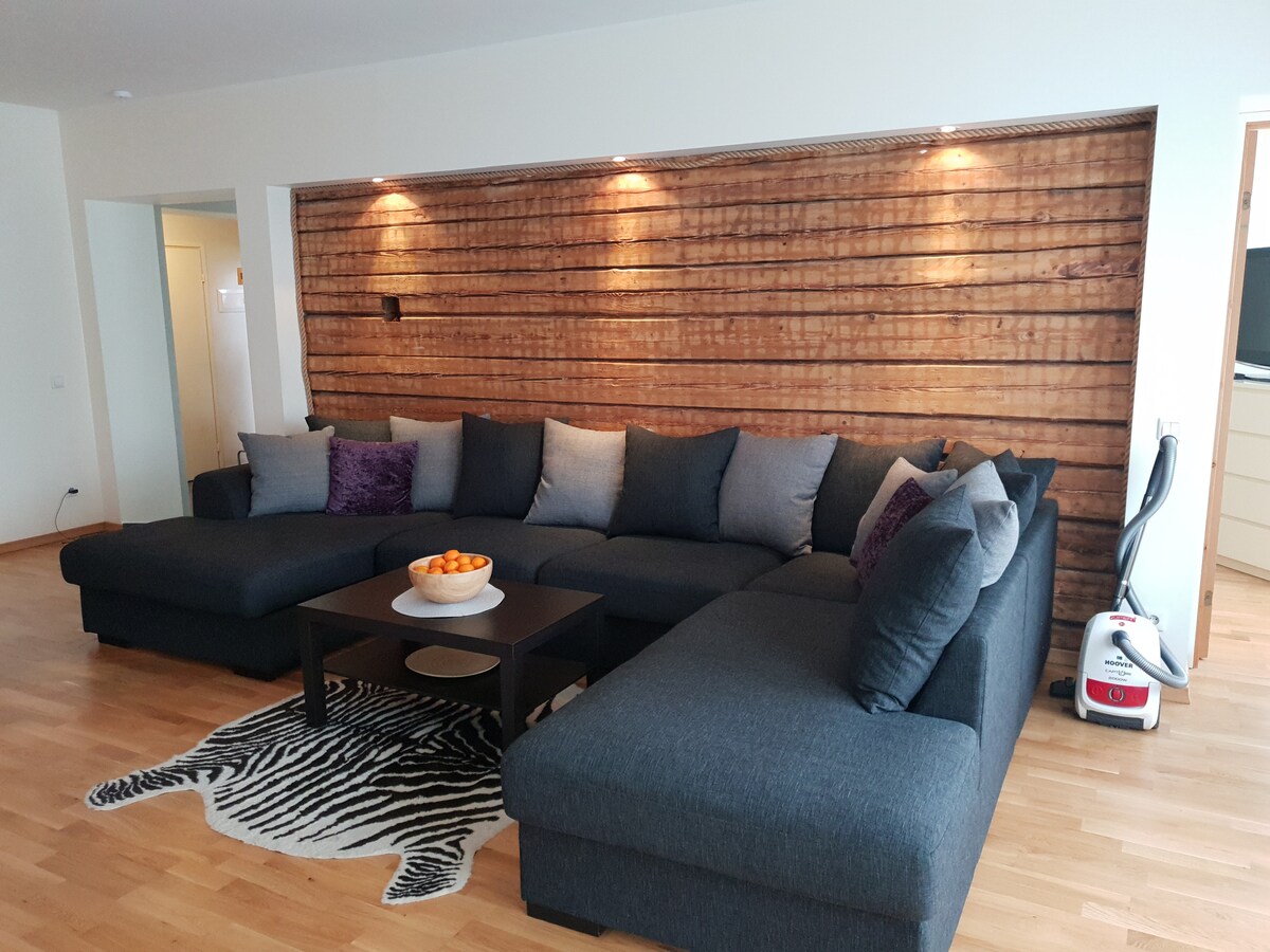 A spacious living area features a large, dark grey sectional sofa adorned with assorted cushions in calming tones. A low black coffee table sits at the center, holding a bowl of fresh oranges. Soft lighting highlights a wooden accent wall behind the seating arrangement.