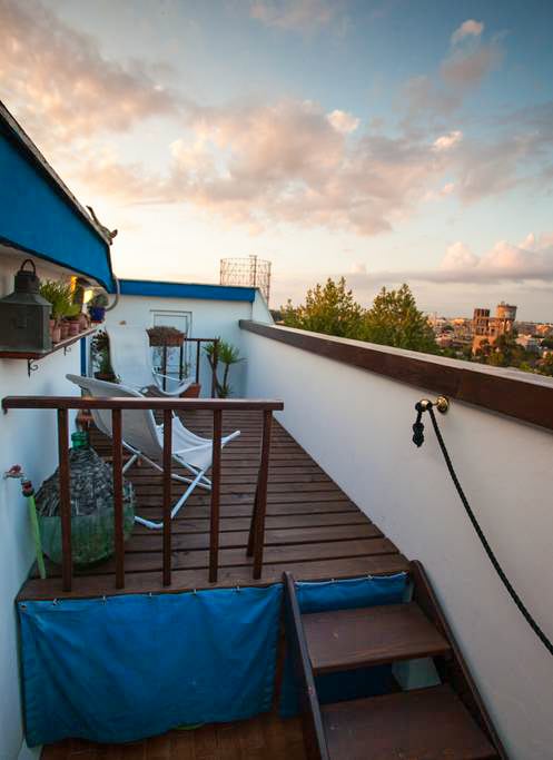 An outdoor terrace is presented with wooden flooring and surrounded by potted plants. A lounge chair and a small table invite relaxation. A staircase provides access, while views of the skyline and distant scenery are framed by the terrace’s protective railing.