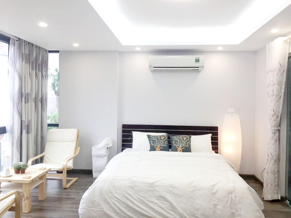 A bright master bedroom features a spacious bed with white linens and decorative pillows. A sitting area is provided by a light-colored chair and a small wooden table. Natural light filters in through large windows, accompanied by minimalist decor and air conditioning unit.