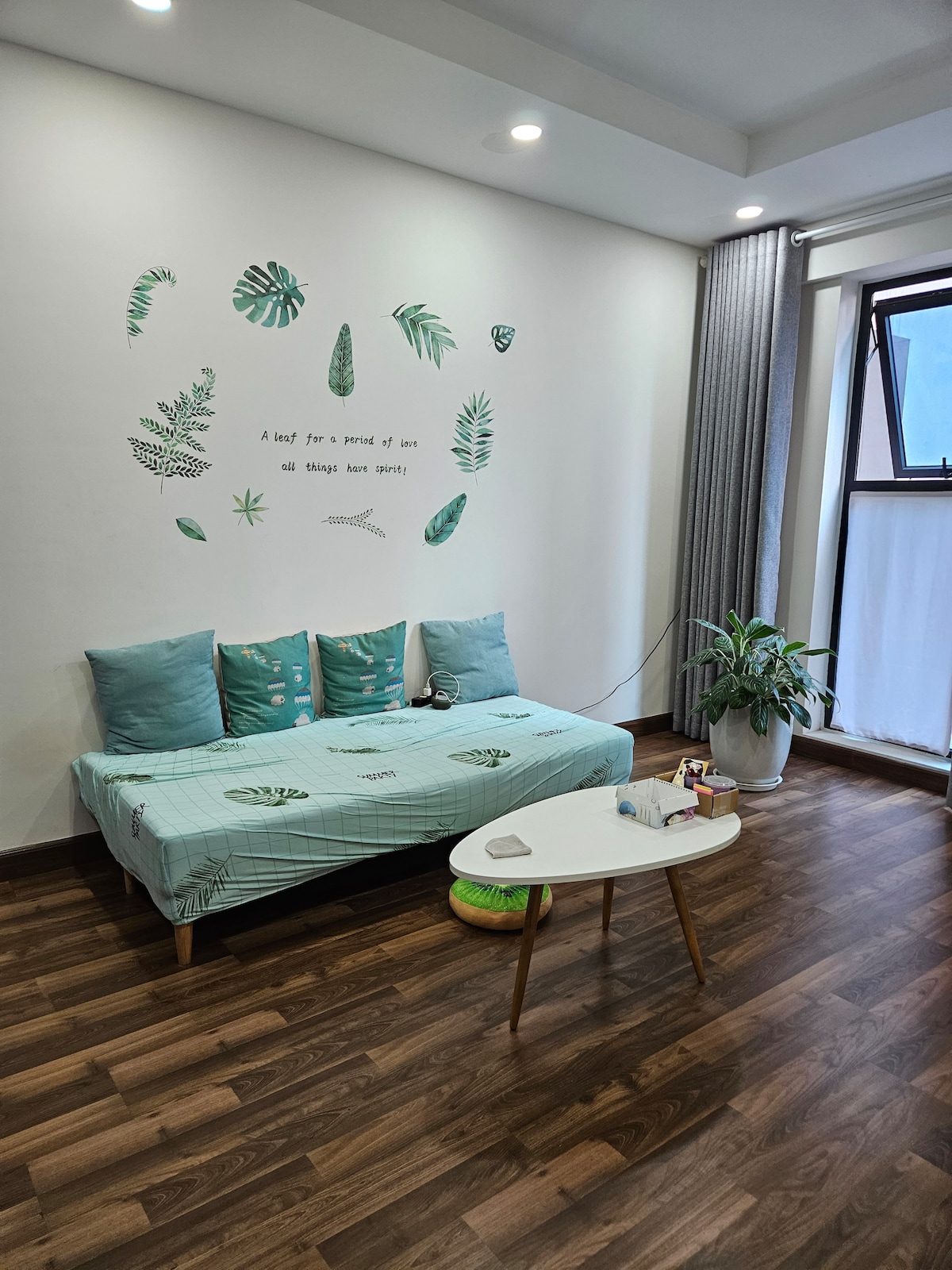 A stylish seating area is featured, equipped with a light blue sofa adorned with decorative pillows. A small coffee table sits nearby, complemented by a houseplant. The wall is decorated with green leaf designs and a motivational quote, enhancing the inviting atmosphere.