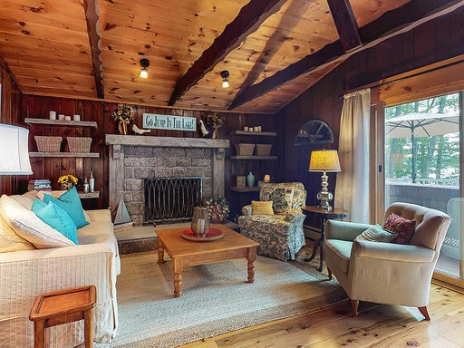 The inviting living room features a large stone wood-burning fireplace as the focal point, surrounded by wooden beams and warm tones. A comfortable sofa and armchair provide relaxing seating options, complemented by a cozy coffee table and soft area rug, creating a welcoming atmosphere.