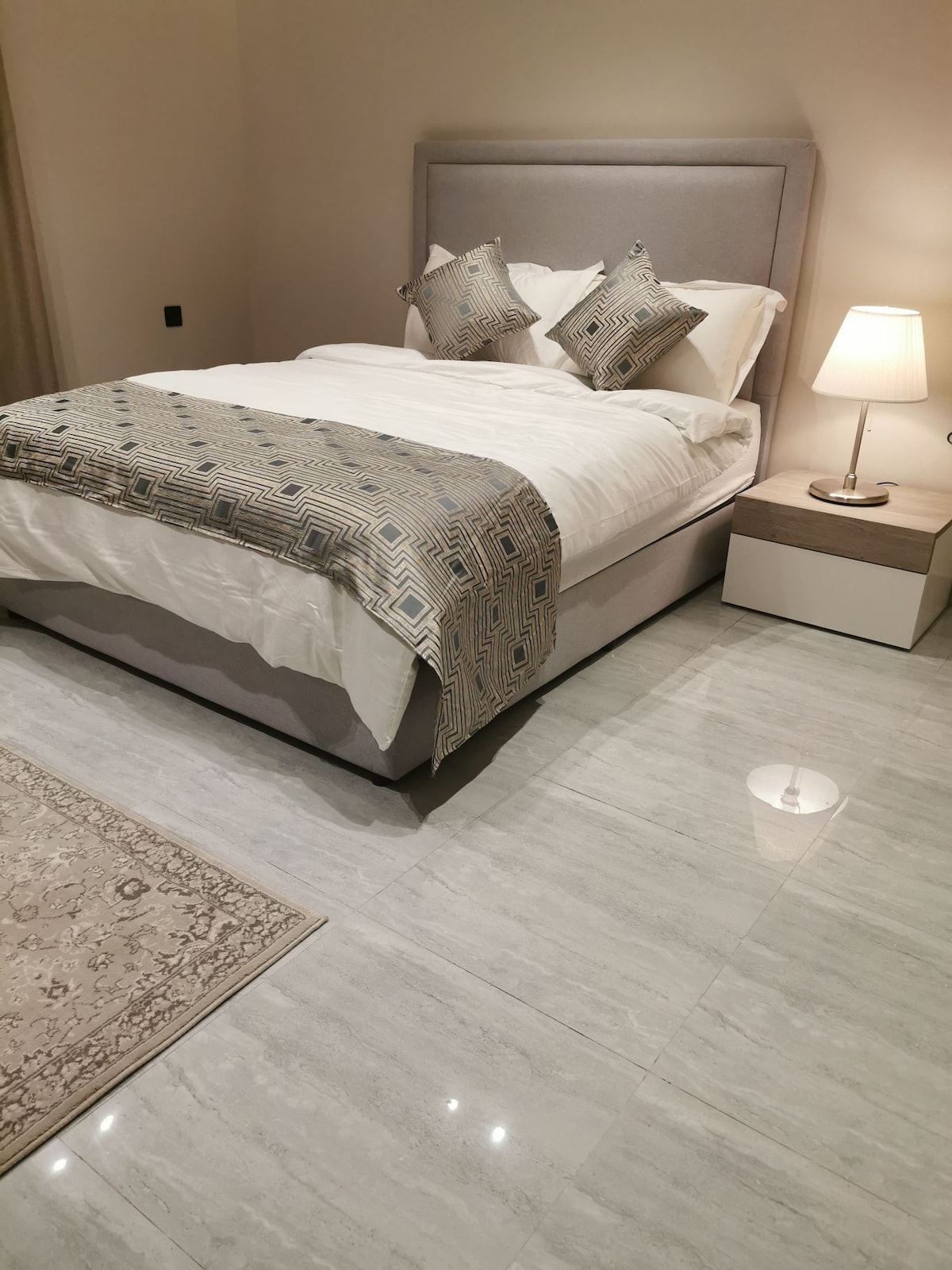 A spacious bedroom features a light-colored upholstered bed with patterned decorative cushions. A bedside lamp provides soft lighting, and a small nightstand sits beside the bed. Polished marble flooring enhances the room's elegance, accompanied by a subtle area rug.