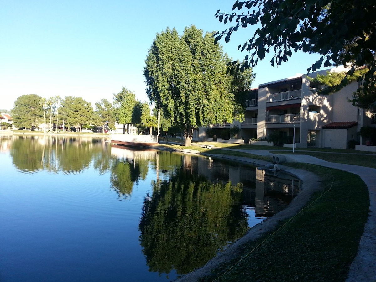 A serene lake reflects the clear blue sky and surrounding greenery. The shoreline features a walking path, with trees providing shade along the route. Condominiums are visible in the background, contributing to the tranquil atmosphere of the area.