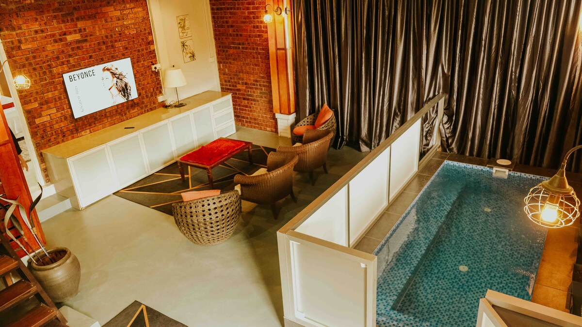 A spacious studio loft features a cozy seating area with two woven armchairs and a low coffee table. A television is mounted on the exposed brick wall. Adjacent, a private pool is visible, enclosed by a stylish partition, adding a unique touch to the living space.