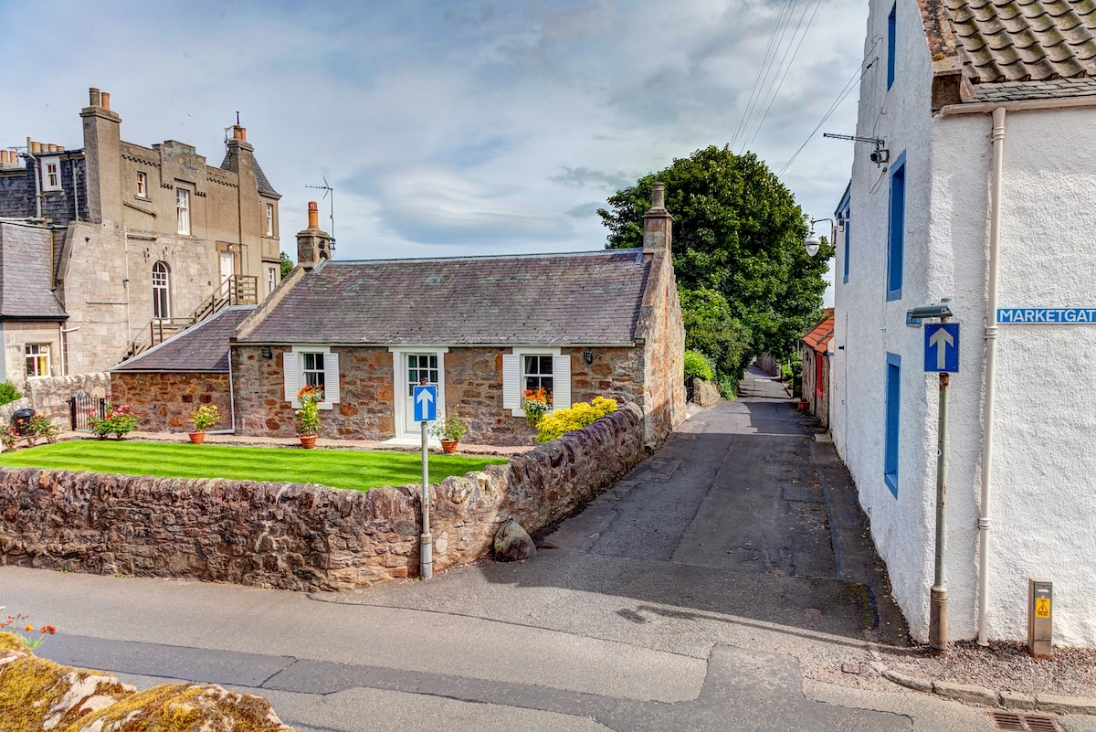 A charming cottage is framed by a well-maintained green lawn and vibrant flowers. The stone structure features traditional architecture and is positioned at a quiet intersection. A nearby street sign indicates the direction to Marketgate, emphasizing the welcoming village setting.