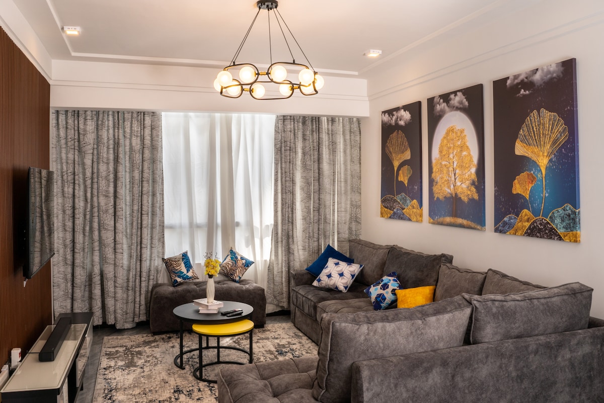 A modern living room features a spacious gray sectional sofa adorned with patterned cushions. A glass coffee table with a yellow top is positioned centrally. Large windows are draped with light-gray curtains, allowing natural light to filter through. Colorful wall art complements the space.