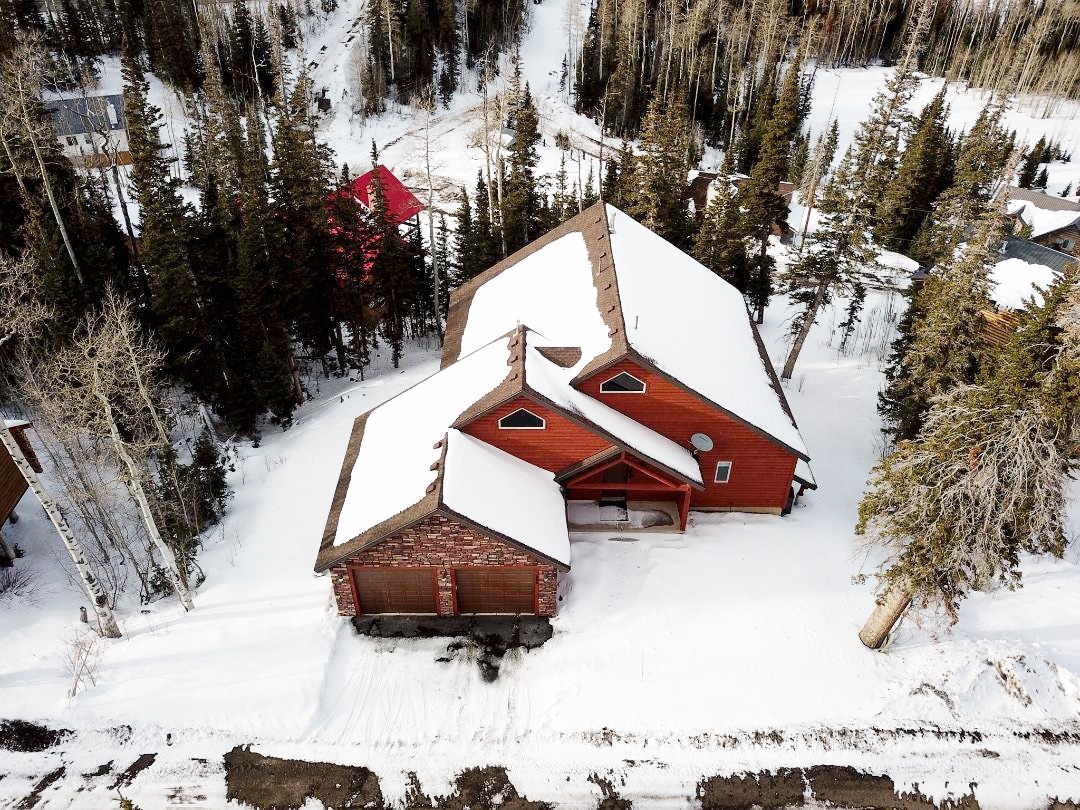 Aerial view of the cabin reveals a spacious structure with a sloped roof, surrounded by snow-covered terrain and tall evergreen trees. The red wood exterior contrasts with the white snow, and a driveway leads to the attached garage, providing a sense of access and warmth in a winter landscape.