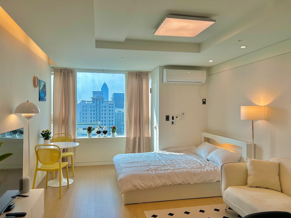 A bright and airy space features a spacious bed covered with fresh linens, complemented by a cozy sofa. A large window allows ample natural light and showcases city views. Yellow chairs and small plants add a touch of color to the modern decor.