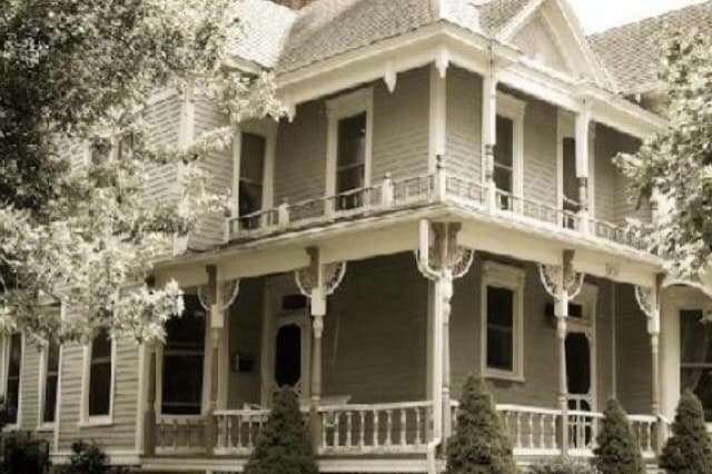 The exterior of the Victorian home showcases its original architectural features, including ornate trim and a wraparound porch. Tall trees frame the house, providing shade and a sense of seclusion, while neatly arranged shrubbery complements the property.