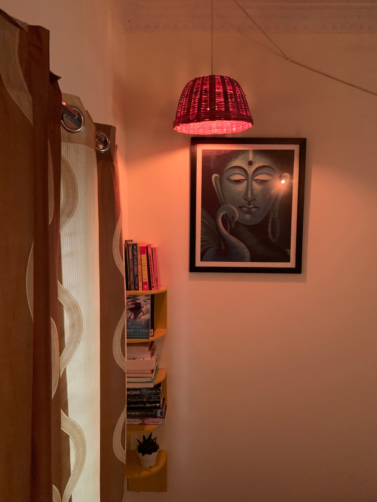 A cozy corner is illuminated by a red pendant light, casting a warm glow. A framed artwork is mounted on the wall, while a small bookshelf holds a collection of books and a potted plant. Soft curtains frame the space, enhancing a relaxing atmosphere.