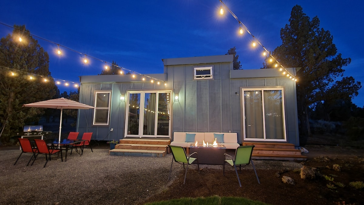 A tiny house is illuminated by string lights at dusk, creating a cozy outdoor atmosphere. The front porch features a fire pit surrounded by colorful chairs and a dining table with an umbrella, enhancing a welcoming space for relaxation.