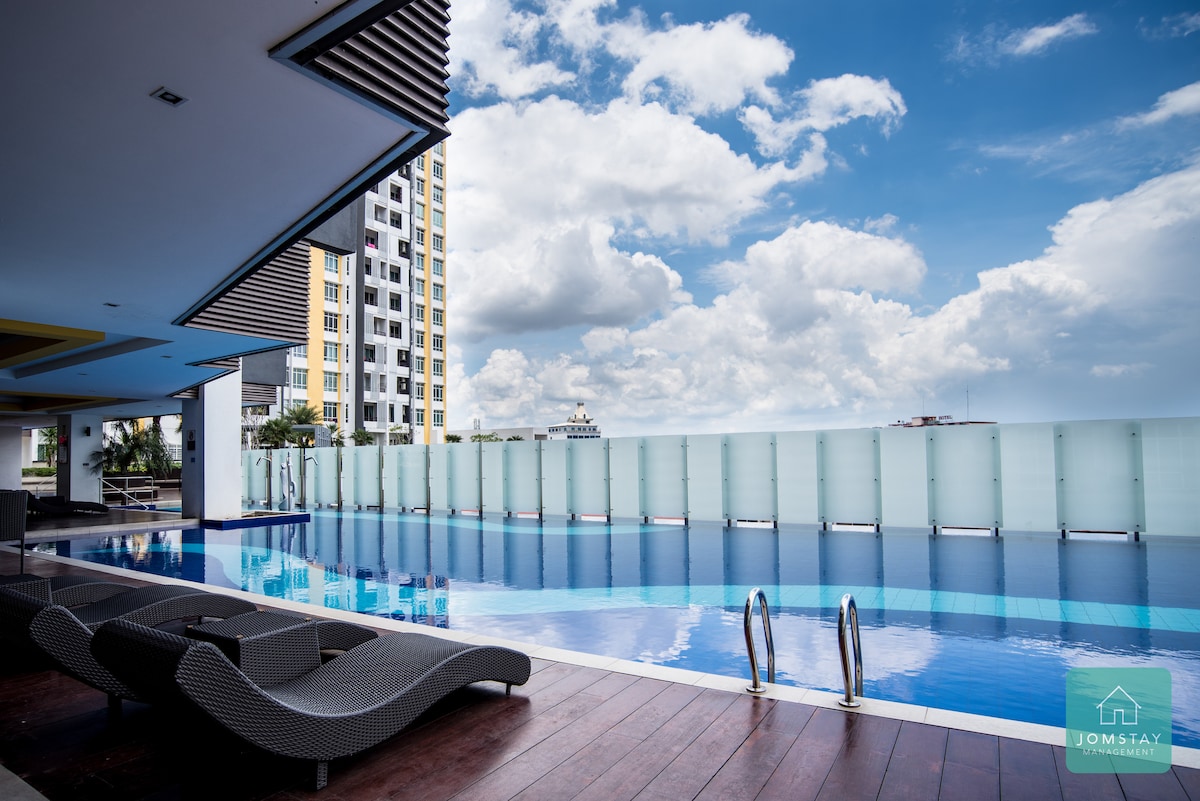 An infinity pool reflects the sky and surrounding buildings. Lounge chairs are positioned alongside the pool, offering a space for relaxation. The area is framed by a contemporary architectural design, and clear glass panels enhance the view of the horizon.
