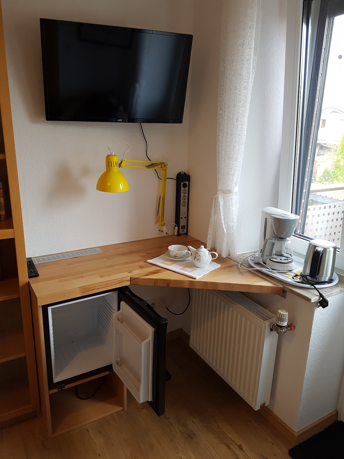 A compact corner desk is shown, featuring a small refrigerator and a coffee machine. A bright yellow lamp provides illumination. A television is mounted on the wall above, while a tray with two cups is placed on the desk. Natural light enters through the window.